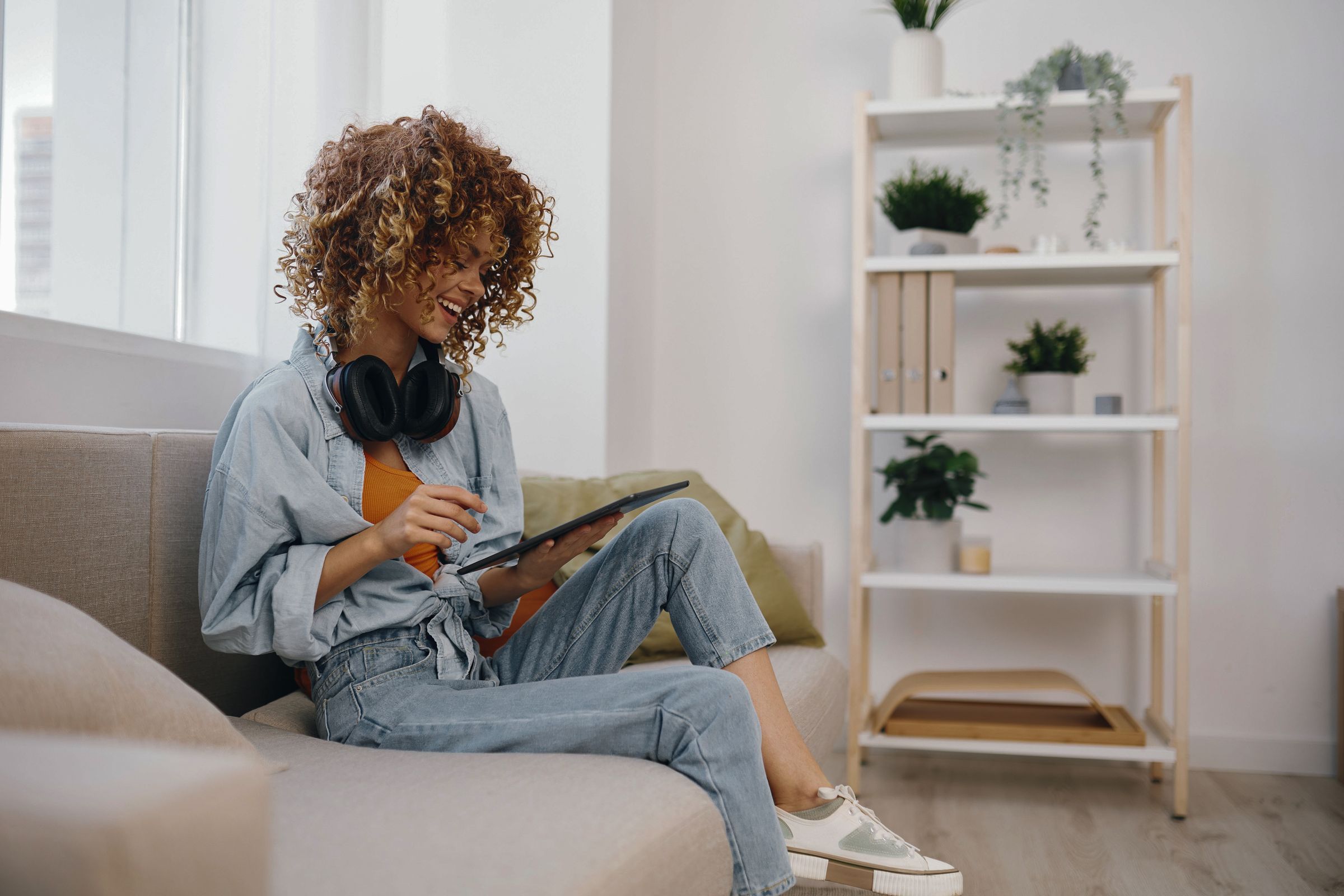 Happy woman holding a tablet PC and listening to music with headphones while relaxing on a cozy couch in her home