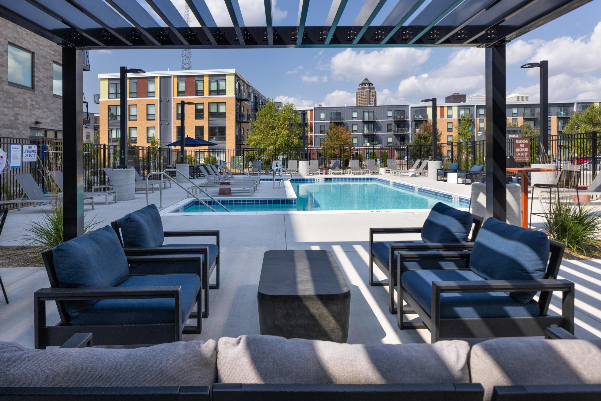 The Meridian modern apartment pool area with lounge seating under a pergola, surrounded by buildings and trees.