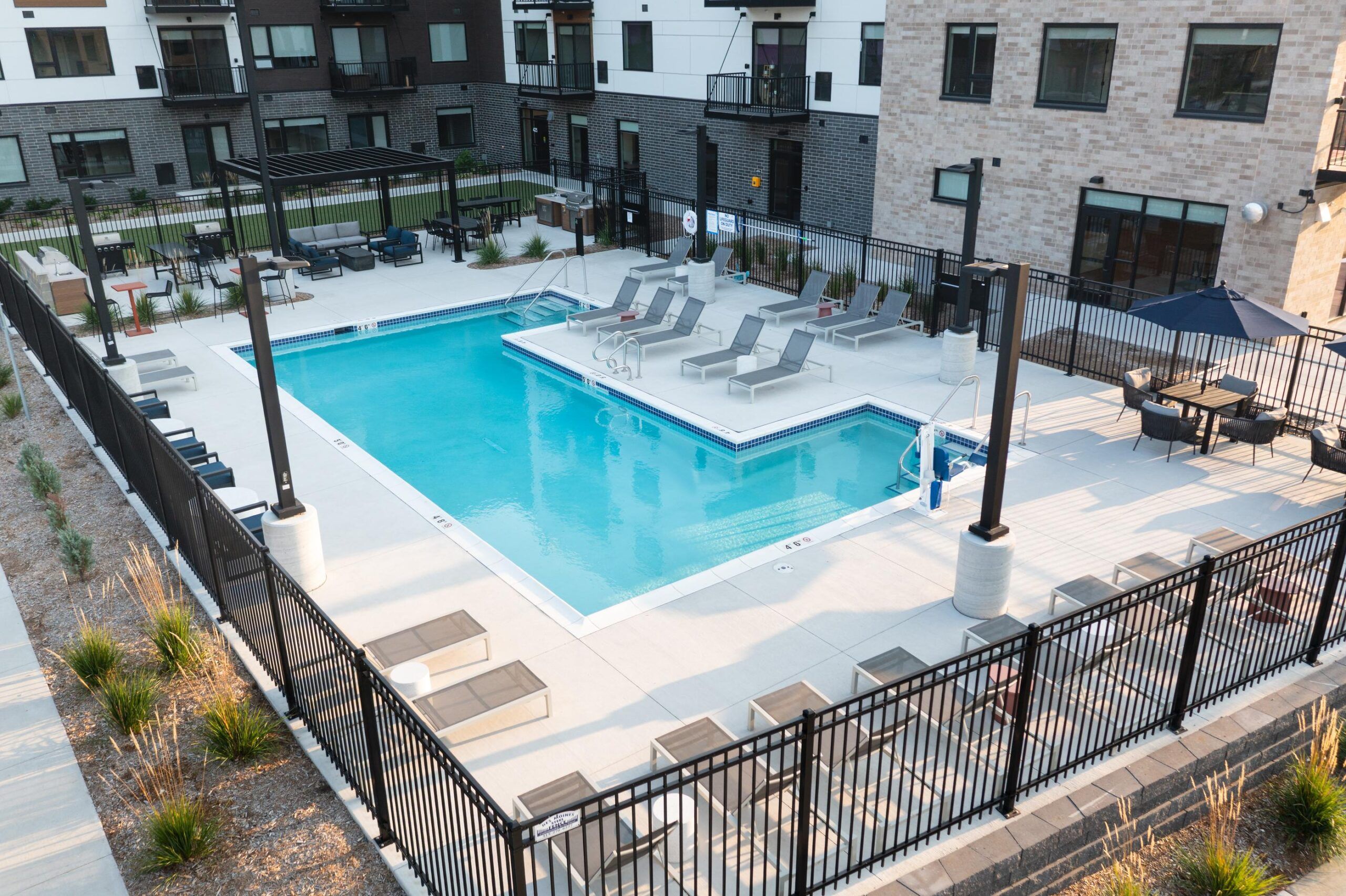 The Meridian fenced-in outdoor apartment pool area with lounge chairs, umbrellas, and nearby modern buildings.