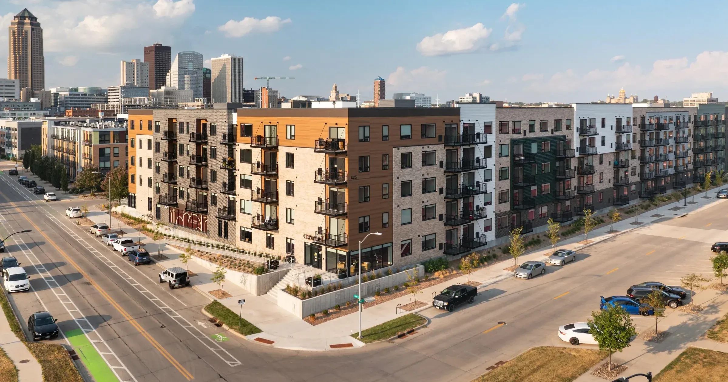 Four-story modern apartment buildings on a city street with cars, trees, and skyscrapers in the background.